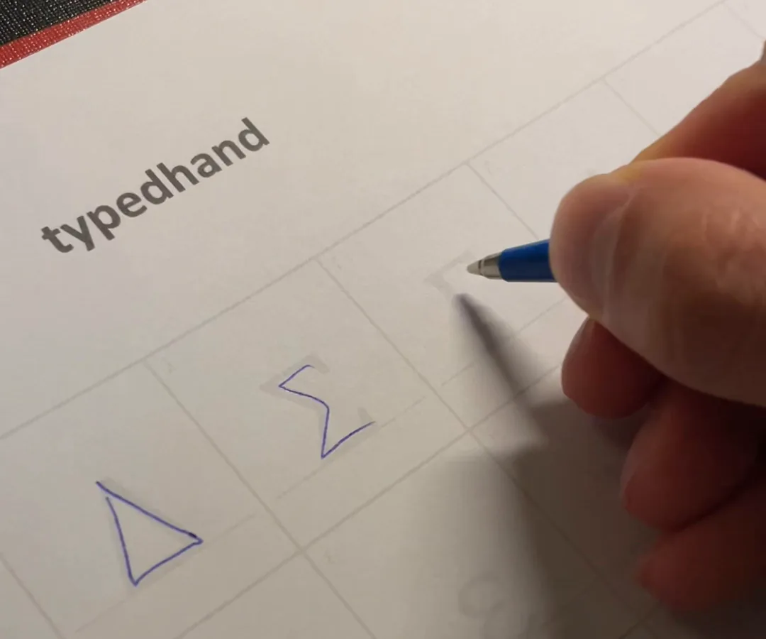 Eine Hand schreibt mit blauem Kugelschreiber die griechischen Großbuchstaben Delta und Sigma in die Kästchen des TypedHand Mathebogens.
